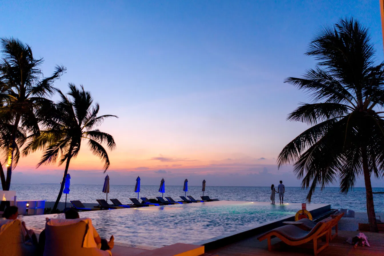 Couple silhouetted at the edge of an infinity pool at dusk, lounge chairs and umbrellas along the horizon