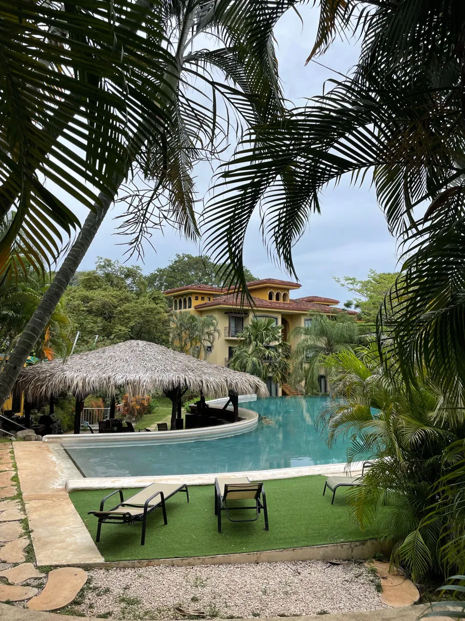 Curved lazy-river pool with thatched palapa and yellow Mediterranean villa